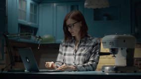 Young 30s woman in glasses working on laptop from home on blue kitchen at night. Business woman talking online by video call working from home. 