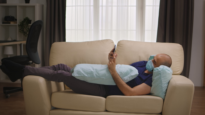 Bored man with protection mask laying on sofa browsing on smartphone during coronavirus self isolation. | Shutterstock HD Video #1053265097