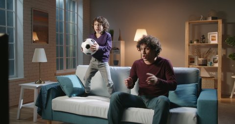 Funny asian male siblings with curly hair spending time together, watching soccer game on tv and emotionally reacting. Authentic father and son enjoying their hobby - family time concept 4k footage