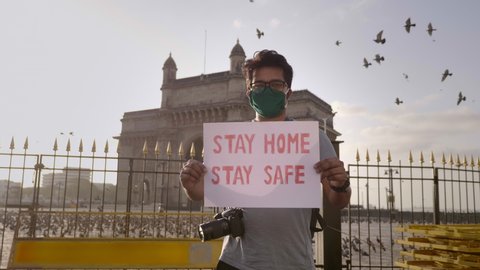 A young man wearing face mask standing and holding a placard with message 'Stay home stay safe' during city lockdown amid coronavirus in front of Gateway of India with pigeons flying in back.