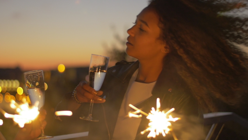Joyful friends enjoying sparklers, toasting and clinking flutes of champagne. Two young cheerful women having fun together at night in the modern urban city.