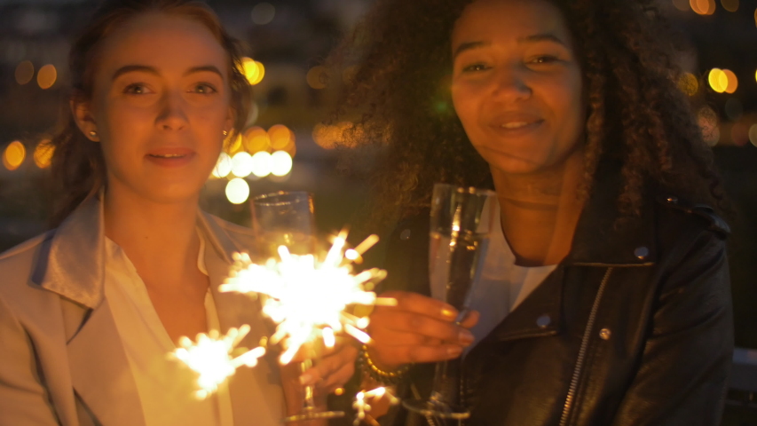 Portrait of happy millennials waving sparks while celebrating a festive outside. Two easy-going friends hanging out at the party with urban city view in background.