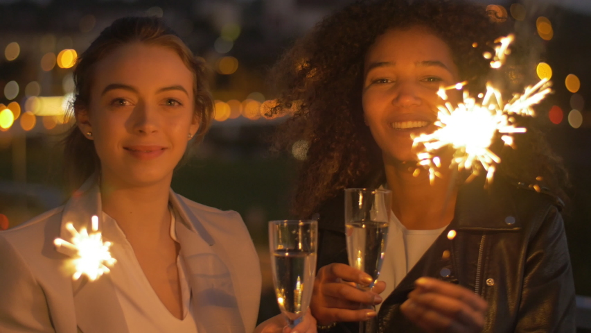Portrait of joyful friends enjoying sparklers and clinking flutes of champagne. Two young cheerful women having fun together at night in the modern urban city.