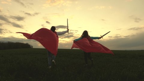 girls in red cloaks run with swords in hand across field playing medieval knights. children fight with toy sword. children play knights. happy childhood concept. healthy young girls play super heroes