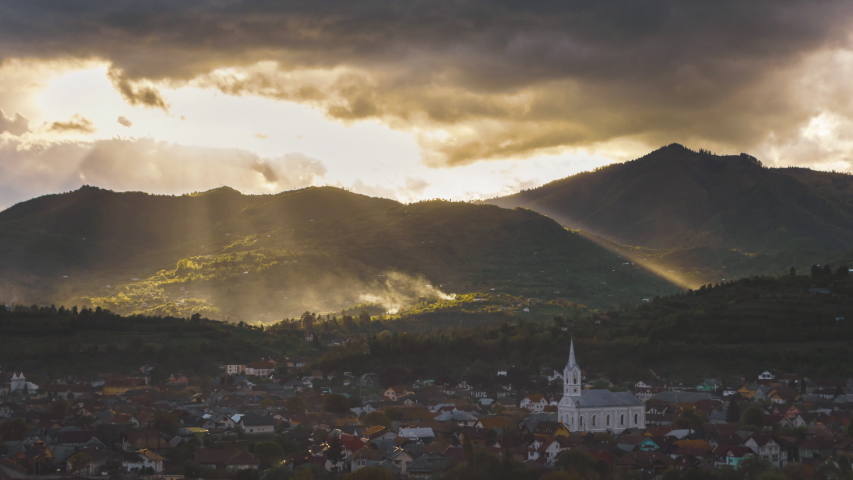 Epic rays and beams of sunlight through the clouds and the rain, rural landscape with church and buildings under the mountains 4k timelapse