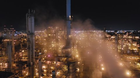 Drone flight over the territory of huge petroleum refinery. Clouds of smoke rise from numerous factory pipes. All constructions are illuminated by factory lighting. The sky is totally black. Aerial,4K