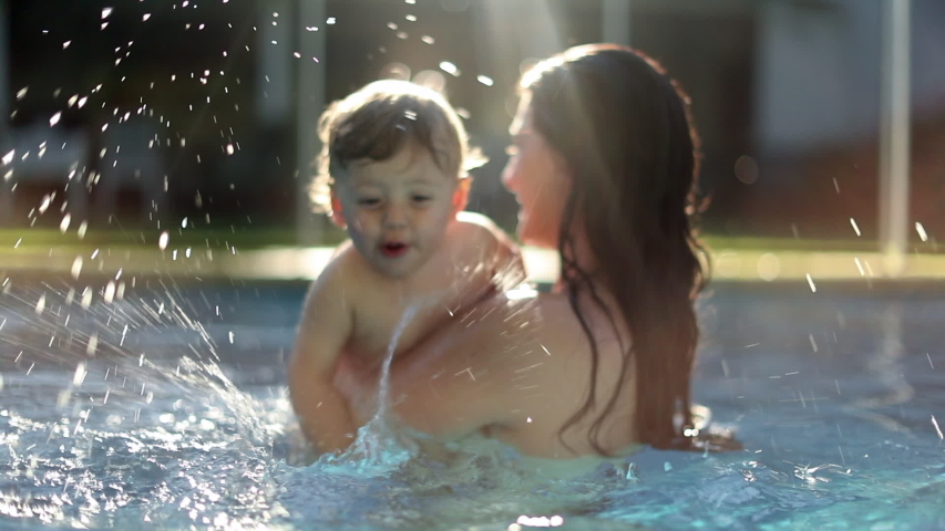 Happy toddler at the swimming pool splashing water in slow-motion 120fps. Mother holding infant son.