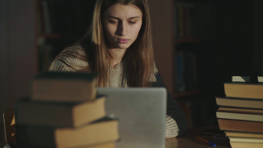 Pretty schoolgirl wears glasses and notes from laptop at desk with books in a library at late time. 4K