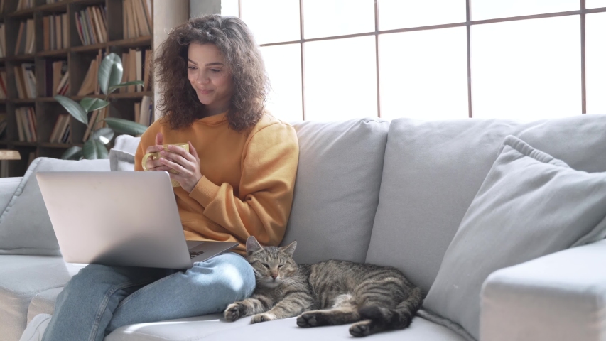 Happy latin teen girl looking at laptop laugh relax with cat on couch at home. Smiling young hispanic woman watching movie holding computer having fun with pet drinking tea sit on sofa in living room. Royalty-Free Stock Footage #1053275708