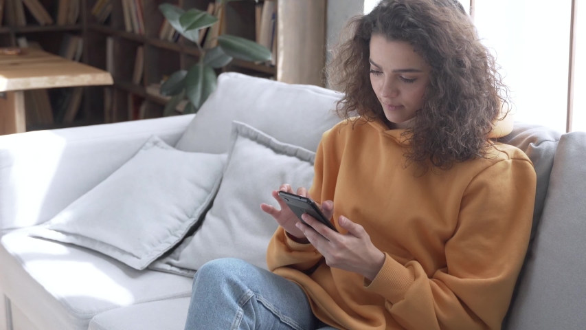 Happy millennial hispanic teen girl checking social media holding smartphone at home. Smiling young latin woman using mobile phone app playing game, shopping online, ordering delivery relax on sofa. Royalty-Free Stock Footage #1053275732