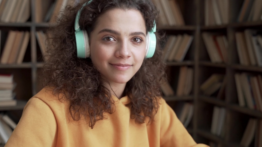 Happy latin teen girl school college student wear headphones look at camera distance learn online on laptop computer in library at home. Zoom video call remote courses, education webinars. Portrait. Royalty-Free Stock Footage #1053275735