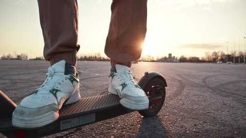 Shot on legs young man in white sneakers ride on electric mobile scooter outside parking space at sunset. Beautiful scenery, nature. Eco-friendly transportation.