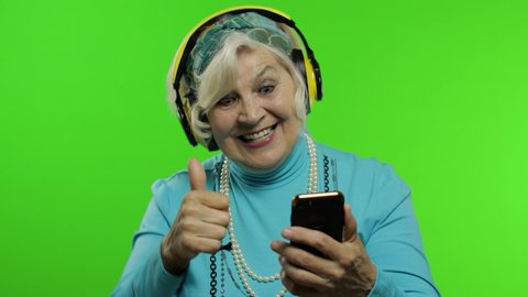 Elderly style granny caucasian mature woman in headphones dancing, listen music from smartphone app on chroma key background. Senior old grandmother in fashion clothing celebrates