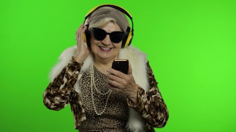 Elderly style granny caucasian mature woman in sunglasses and headphones dancing, listen music from smartphone app on chroma key background. Senior old grandmother in fashion clothing celebrates