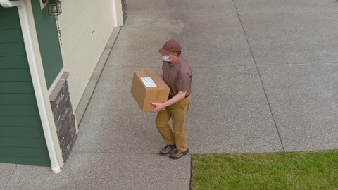 Mail courier wearing a medical face mask delivers a mask delivers a package to a home while practicing social distancing; COVID-19 pandemic theme