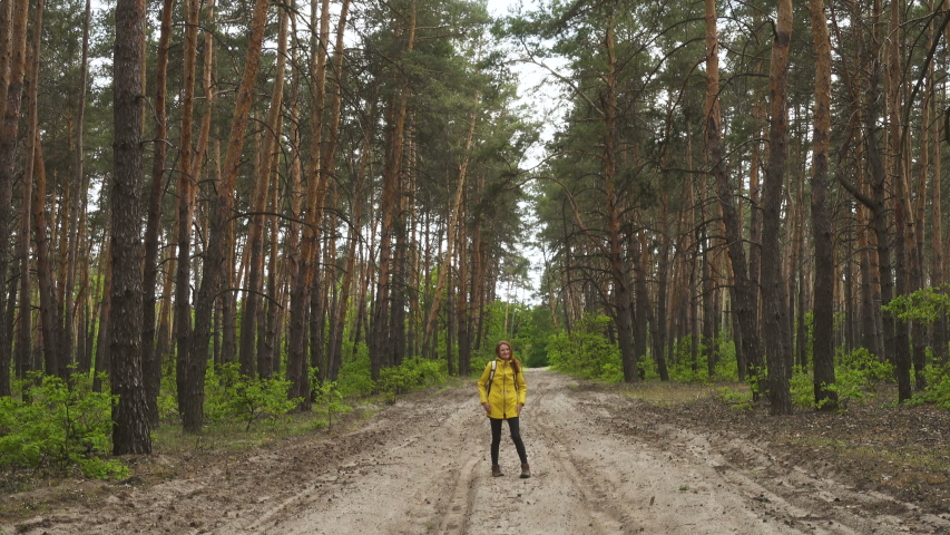 smiling hiker girl wearing yellow jacket with the backpack walking by the pine forest road 
