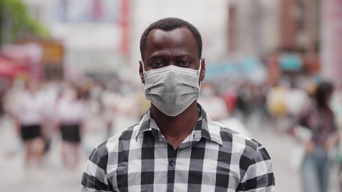 Slow motion of one black African man wear face mask looking at camera smile in the eye at urban street with crowd of people walking in blurred background coronavirus Covid-19 pandemic outbreak