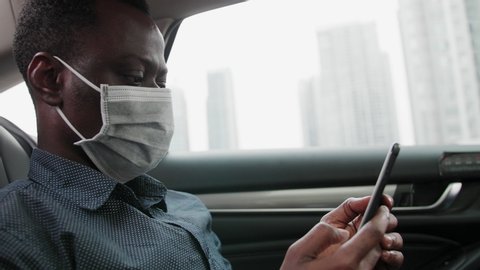 Side view of one black businessman wear mask traveling in the car using mobile phone looking at phone in hand during pandemic of coronavirus Covid-19