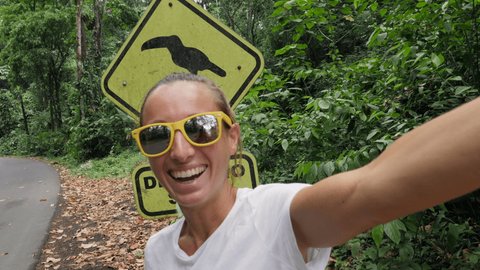 woman taking selfie video with Slow down Toucan crossing sign inCosta Rica. People travel sharing online video chatting. Girl exploring tropical country 