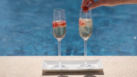 Close up of two glasses with champagne or prosecco with red berries on swimming pool or sea background. Sparkling white wine on poolside, blurred backdrop. Female hand putting raspberry in glasses.