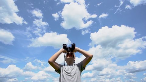 Cute white young kid looking through old black vintage binoculars at something interesting far away in distance. Boy stands isolated on sunny summer blight blue sky and white fluffy clouds background.