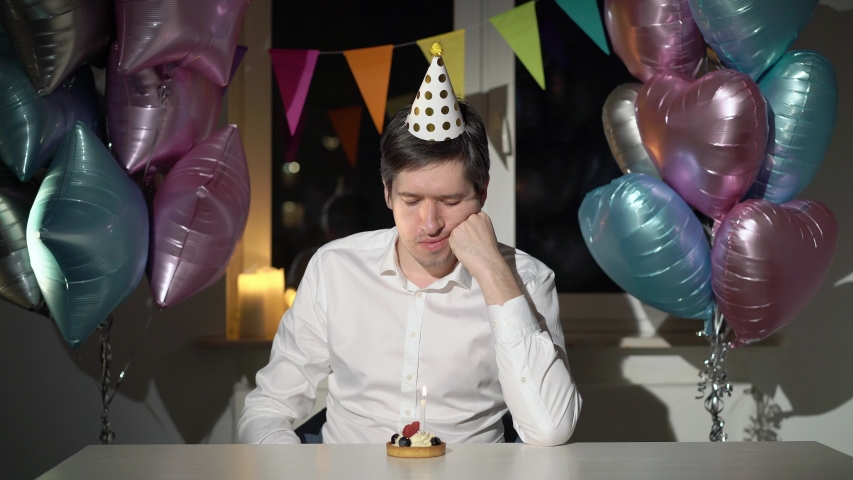 Young man celebrating birthday alone. Sad bored and tired male bday with party cake and blowing candle, balloons