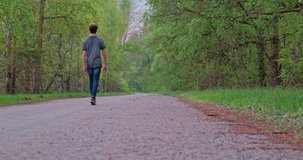 Rear view to the Guy  strolling on the nature. Young slim man is walking alone on the road. Peaceful teenager walks through outdoors.   Human feel a freedom walking alone on the asphalt road. 4k video