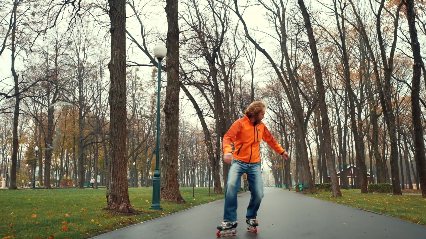 Sporty bearded European male roller skater in orange jacket performs complex and technical movements and ice skates in the alley of the central city park. Healthy lifestyle and active leisure outside.