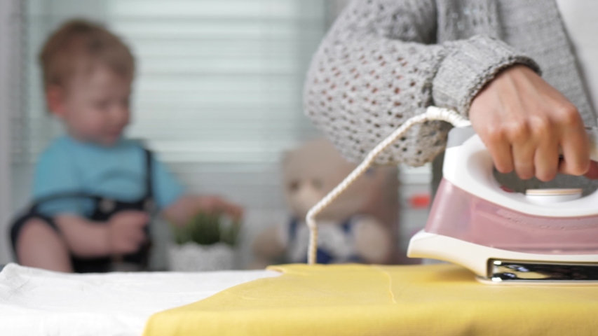 Girl ironing clothes. Female hand ironing yellow dress, on blurred background small child sits on windowsill and plays | Shutterstock HD Video #1053316505