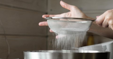 Woman is sifting flour through sieve for baking mixing ingredients in metal bowl, hands closeup view. Preparing flour to baking cake in kitchen at home. Steps cooking baking and confectionery.