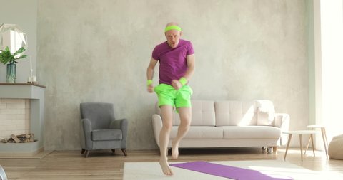 Man is running in place at home making sport fitness exercise, camera moves away from face. Funny portrait of young guy. Sportive humor. Joke, comical, mem, parody, joking behavior. Bright sportswear.