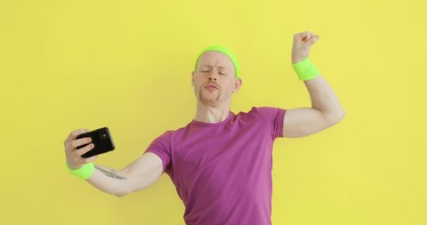 Humorous blogger man is taking picture of his biceps on smartphone on yellow backround. Guy kissing his biceps and boasting showing muscles online. Joke, comical, mem, parody, sportive humor concept.