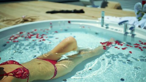 The woman lifts her leg in the jacuzzi decorated with rose petals. slow motion.