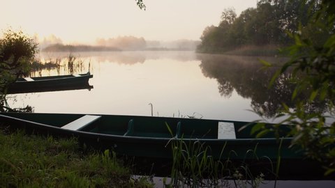Beautiful early morning peaceful countryside landscape. 4k video footage of old wooden rowboats in foreground, foggy water of small river, misty horizon line and dark trees growing at shores.