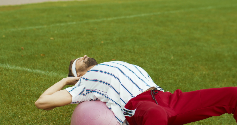 Handsome funny vintage sportsman doing abs exercises on fitness ball outside. Caucasian old-fashioned male athlete having workout with gym ball outdoors alone on stadium. Sportsman concept