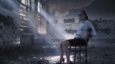 Kidnapped woman looks around in abandoned building while sitting tied to chair in captivity.