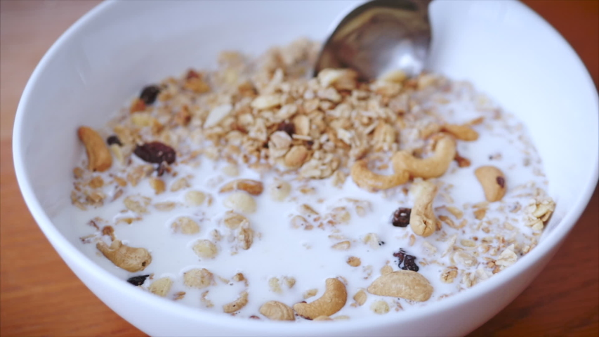 Close-up shot of a female hand stirring a spoon of cereal in a breakfast plate. The concept of breakfast, a healthy diet, granola with cashew nuts and slices of raspberries and raisins. | Shutterstock HD Video #1053326816