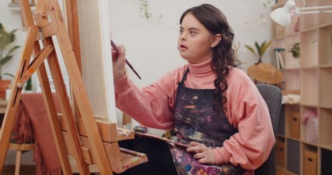Young talented teenager wearing artists apron painting while sitting in front of molbert at her room. Pretty girl with genetic disorder holding palette on her knees creating picture