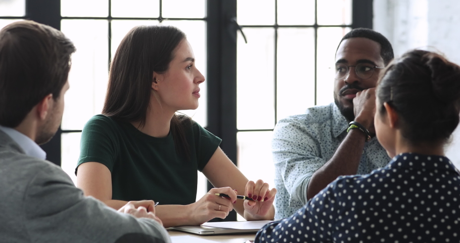 Young female team leader sitting at table with mixed race colleagues, discussing working issues at negotiations meeting. Skilled millennial diverse employees developing startup marketing strategy. Royalty-Free Stock Footage #1053331487