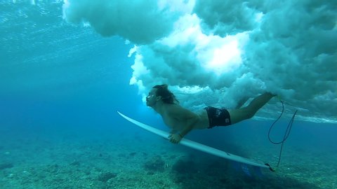 SLOW MOTION, UNDERWATER: Fit Caucasian surfer does a duck dive under a rushing barrel wave. Cinematic shot of a male surfer duck diving into the beautiful blue ocean water and under an incoming wave.