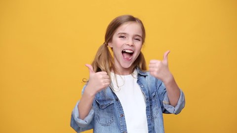 Little blonde kid teen girl 12-13 years old in denim jacket white t-shirt posing isolated on yellow background studio. People childhood lifestyle concept Looking at camera laughing showing thumbs up
