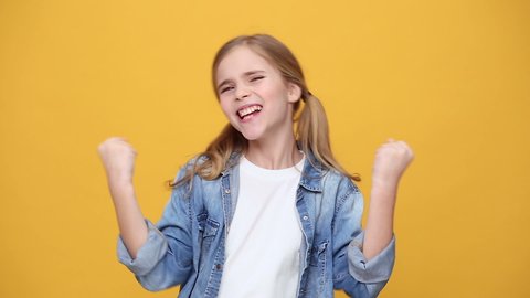 Little fun happy blonde kid teen girl 12-13 years old in denim jacket white t-shirt posing clenching fists doing winner gesture say Yes isolated on yellow background studio. People lifestyle concept