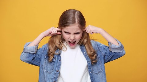 Little angry blonde kid teen teenager girl 12-13 years old in denim jacket white t-shirt isolated on yellow background studio. People childhood lifestyle concept cover ears with hands scream scolding