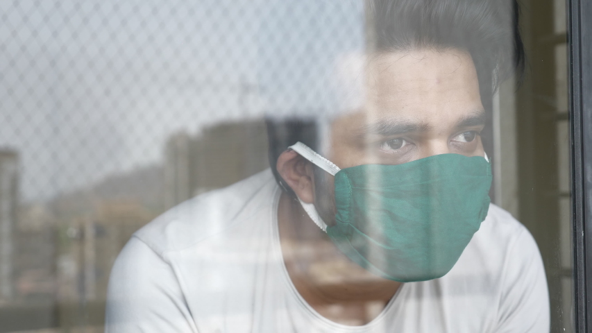 A shot of a young man through the glass window is wearing a green protective face mask and looking out. A distressed or worried patient is quarantined during Coronavirus or COVID 19 epidemic   | Shutterstock HD Video #1053334340