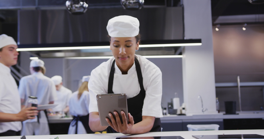 Professional African American female chef wearing chefs whites in a restaurant kitchen, using a tablet, with colleagues working in the background in slow motion Royalty-Free Stock Footage #1053335699