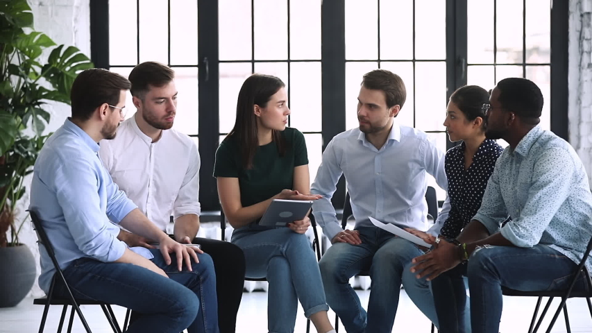 Focused young mixed race business people sitting on chairs in circle, discussing working issues in relaxed atmosphere. Skilled female psychologist trainer counseling mentoring diverse team in office. Royalty-Free Stock Footage #1053336191