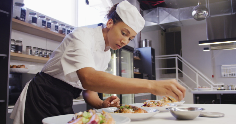 A professional African American female chef wearing chefs whites in a restaurant kitchen, putting food on a plate in slow motion Royalty-Free Stock Footage #1053336356