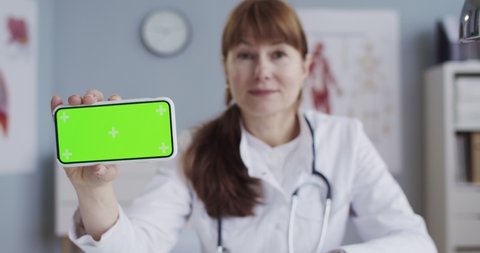 Pretty joyful Caucasian female doctor in white gown holding smartphone with green screen while sitting in cabinet. Beautiful happy physician showing mobile phone with chroma key with tracking motion