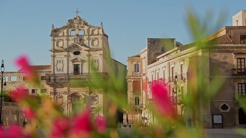 Camera movement through the bright flowers and the catholic church of Saint Lucia alla Badia in the empty square of Ortigia island in province of Syracuse in Italy