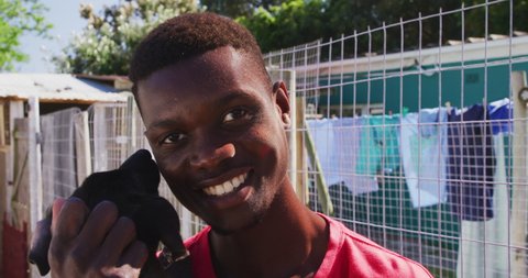 Portrait of an African American male volunteer wearing red tshirt at an animal shelter, holding a rescued dog in his arms, smiling and looking at camera, on a sunny day in slow motion